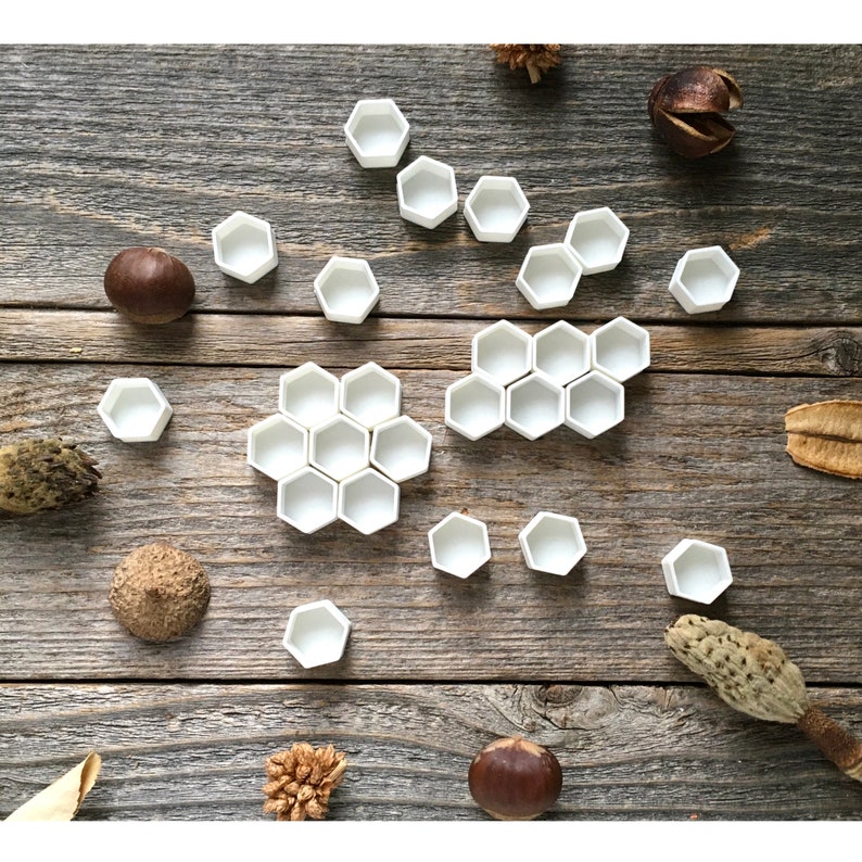 Może przedstawiać: Białe sześciokątne pojemniki ceramiczne ułożone w kształcie plastra miodu na drewnianej powierzchni. Pojemniki są r&oacute;żnych rozmiar&oacute;w i rozrzucone po powierzchni.