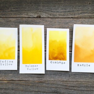 May include: Four rectangular white paper swatches featuring watercolor paint gradients in shades of yellow and orange. The swatches are labeled with black text reading Indian Yellow, Dalamar Yellow, Gamboge, and Rutile, arranged in a horizontal row.