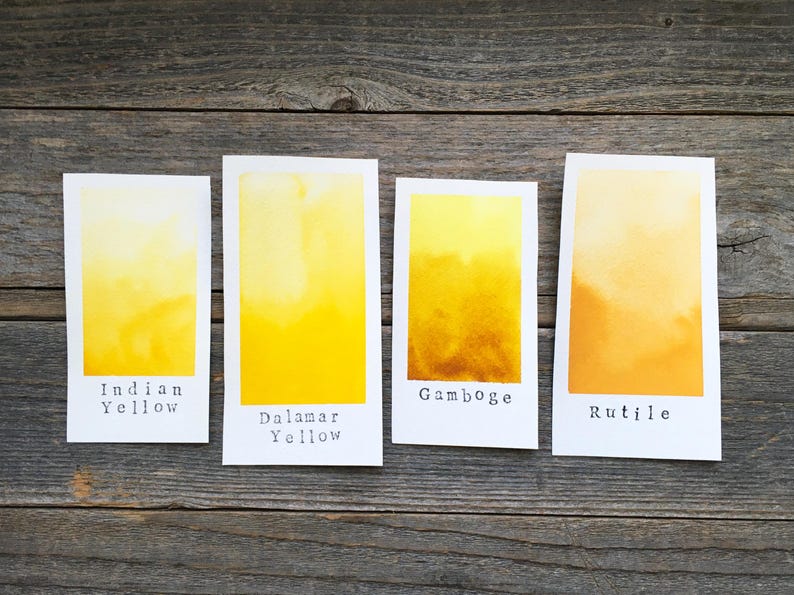 May include: Four rectangular white paper swatches featuring watercolor paint gradients in shades of yellow and orange. The swatches are labeled with black text reading Indian Yellow, Dalamar Yellow, Gamboge, and Rutile, arranged in a horizontal row.