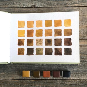 May include: A watercolor swatch chart on white paper featuring twenty-four square samples arranged in four rows, displaying a gradient of earth tones from light yellow to dark brown, accompanied by five small rectangular paint pans below.