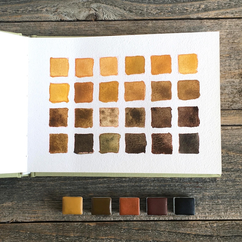 May include: A watercolor swatch chart on white paper featuring twenty-four square samples arranged in four rows, displaying a gradient of earth tones from light yellow to dark brown, accompanied by five small rectangular paint pans below.