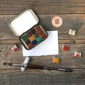 Puede incluir: Un juego de pintura de acuarela con una variedad de colores en una lata de metal, un pincel y varios bloques de pintura de acuarela cuadrados pequeños. Las palabras "POEMS ABOUT YOU" están impresas en varios bloques rectangulares pequeños y una pegatina redonda.