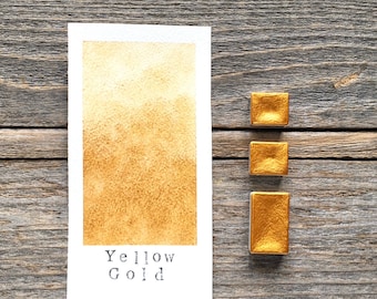 May include: A rectangular white paper swatch featuring a metallic yellow gold watercolor paint sample with the text Yellow Gold printed below it, accompanied by three small rectangular pans of the same metallic yellow gold watercolor paint.