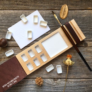 Może przedstawiać: Drewniana paleta do akwareli z brązową sk&oacute;rzaną okładką i wbudowanym uchwytem na pędzle. Paleta ma 10 wgłębień na farby akwarelowe i dużą powierzchnię do mieszania kolor&oacute;w. Paleta jest otwarta, a okładka jest odchylona do tyłu, aby odsłonić wgłębienia. Paleta znajduje się na drewnianej powierzchni z kilkoma małymi elementami dekoracyjnymi.