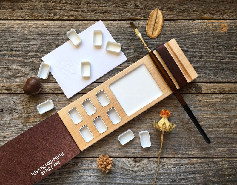 Może przedstawiać: Drewniana paleta do akwareli z brązową sk&oacute;rzaną okładką i wbudowanym uchwytem na pędzle. Paleta ma 10 wgłębień na farby akwarelowe i dużą powierzchnię do mieszania kolor&oacute;w. Paleta jest otwarta, a okładka jest odchylona do tyłu, aby odsłonić wgłębienia. Paleta znajduje się na drewnianej powierzchni z kilkoma małymi elementami dekoracyjnymi.