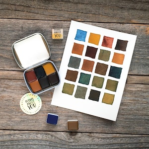May include: A metal watercolor tin containing six paint pans, a white paper swatch card with twenty-four painted squares in various earth tones, a small square stamp reading POEMS ABOUT YOU, and a round sticker with the same text.