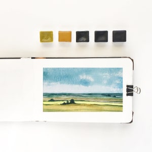 May include: An open sketchbook displaying a watercolor landscape painting of a field under a blue sky with clouds. Above the sketchbook are five square watercolor paint pans in shades of olive green, ochre, and three variations of dark gray and black.