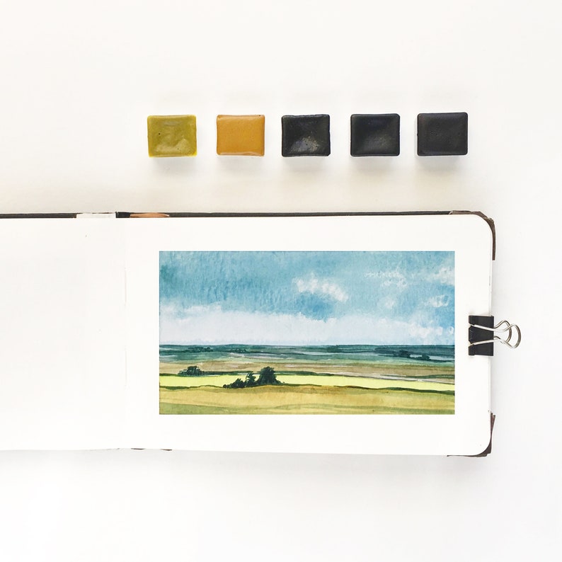 May include: An open sketchbook displaying a watercolor landscape painting of a field under a blue sky with clouds. Above the sketchbook are five square watercolor paint pans in shades of olive green, ochre, and three variations of dark gray and black.
