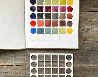 May include: An open sketchbook displaying a grid of painted watercolor swatches in various shades of blue, green, red, brown, and yellow, arranged in circles and squares. Below the book lies a white plastic stencil with matching cutouts.