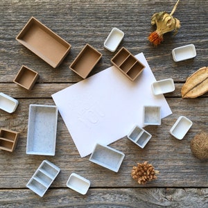 Może przedstawiać: Zbi&oacute;r palet do farb akwarelowych w r&oacute;żnych rozmiarach i kolorach, ułożonych na drewnianej powierzchni. Niekt&oacute;re palety są brązowe, a niekt&oacute;re białe. Wok&oacute;ł palet rozrzucone są r&oacute;wnież suszone kwiaty i inne naturalne elementy.