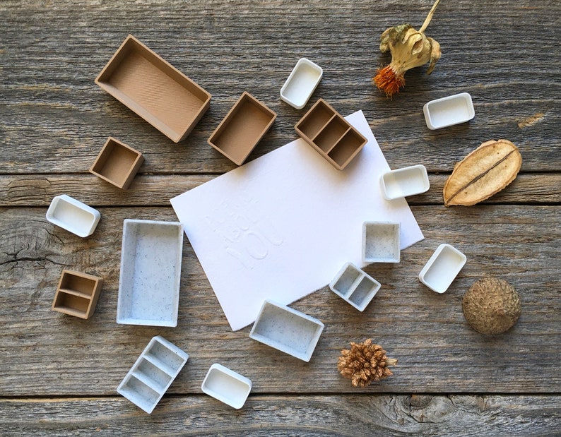 Może przedstawiać: Zbi&oacute;r palet do farb akwarelowych w r&oacute;żnych rozmiarach i kolorach, ułożonych na drewnianej powierzchni. Niekt&oacute;re palety są brązowe, a niekt&oacute;re białe. Wok&oacute;ł palet rozrzucone są r&oacute;wnież suszone kwiaty i inne naturalne elementy.