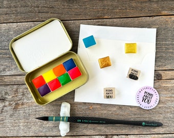 May include: A gold metal tin containing eight rectangular watercolor pans in purple, red, orange, yellow, and blue. Includes a black paintbrush, two wooden blocks and a circular sticker, all featuring the text Poems About You, plus extra paint pans.