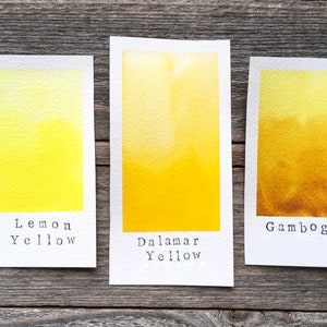 May include: Three rectangular paper swatches featuring watercolor paint samples. The left swatch is labeled Lemon Yellow, the center is labeled Dalamar Yellow, and the right swatch is labeled Gamboge, showing a gradient from light to dark yellow.