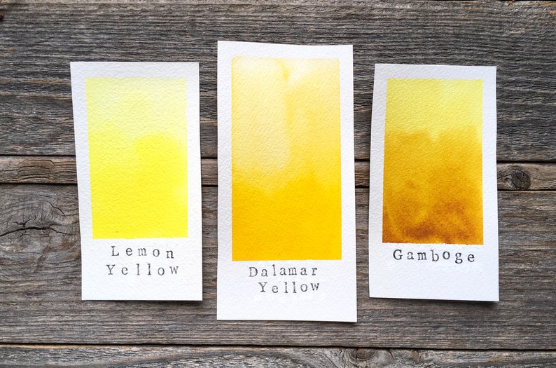 May include: Three rectangular paper swatches featuring watercolor paint samples. The left swatch is labeled Lemon Yellow, the center is labeled Dalamar Yellow, and the right swatch is labeled Gamboge, showing a gradient from light to dark yellow.