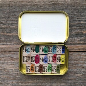 May include: A gold-colored metal tin containing fourteen small rectangular paper scrolls. Each scroll is wrapped in a band printed with the text "POEMS ABOUT YOU" and features a unique watercolor splash in colors including blue, green, pink, and yellow.