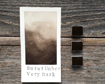 May include: A rectangular watercolour swatch card showing a gradient from light beige to deep brown, labelled "Burnt Umber Very Dark" in black text, accompanied by three small, square, dark brown watercolour paint pans arranged in a vertical column.