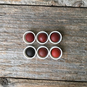 May include: Six small, round objects in varying shades of red and brown, each nestled in a white, circular container. The objects are arranged in two rows of three against a weathered, wooden background.