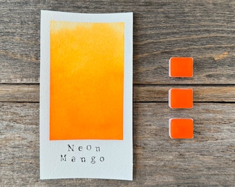 May include: A rectangular white paper swatch featuring a vibrant yellow-to-orange gradient wash with the text Neon Mango printed in black ink at the bottom, accompanied by three small, square, solid orange watercolor paint pans arranged vertically.