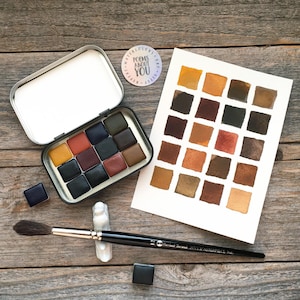 May include: A metal watercolor tin containing twelve rectangular paint pans, a white paper swatch card with twenty-four painted squares in earth tones, a black paintbrush with the text "Borrani-Boriani 102/2 MASTERPIECE Italy", and a round sticker reading "POEMS ABOUT YOU".