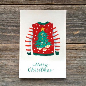 Puede incluir: Una tarjeta de felicitaci&oacute;n rectangular con una ilustraci&oacute;n en acuarela de un su&eacute;ter rojo con lunares blancos, mangas verdes con rayas rojas y un &aacute;rbol de Navidad verde central con una estrella amarilla, bastones de caramelo y regalos, con el texto Merry Christmas.