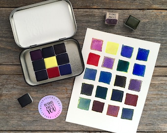 May include: A metal tin containing nine square watercolor pans in shades of purple, yellow, red, and blue. A white paper swatch card displays twenty-four painted squares in various colors. Includes a round sticker and a small stamp labeled POEMS ABOUT YOU.