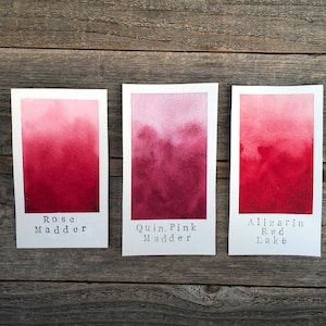 Op de afbeelding: Drie aquarelverfstaaltjes op een houten oppervlak. De staaltjes zijn gelabeld met "Rose Madder", "Quin. Pink Madder" en "Alizarin Red Lake".
