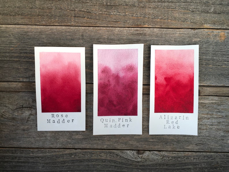Op de afbeelding: Drie aquarelverfstaaltjes op een houten oppervlak. De staaltjes zijn gelabeld met "Rose Madder", "Quin. Pink Madder" en "Alizarin Red Lake".
