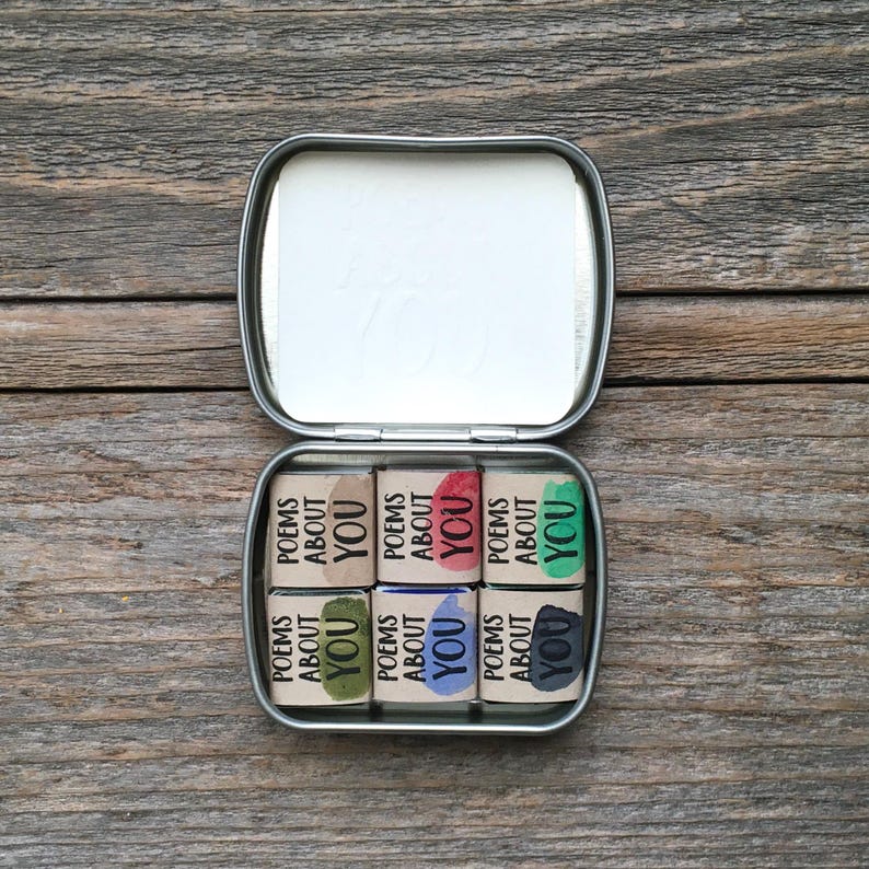 Puede incluir: Una lata rectangular de metal que contiene seis peque&ntilde;os bloques cuadrados, cada uno envuelto en papel color canela con el texto "POEMS ABOUT YOU" impreso en negro. Cada bloque presenta una mancha de tinta de color &uacute;nico en marr&oacute;n, rojo, verde, oliva, azul o gris oscuro.