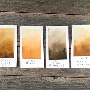 May include: Four rectangular paper swatches displaying watercolor gradients. The labels read Fawn Ocher Deep, Raw Sienna, Raw Umber Greenish, and Cool Ocher Mexico. Each swatch features a smooth transition from a light tone at the top to a darker, saturated hue at the bottom, showcasing distinct earth-toned pigments.