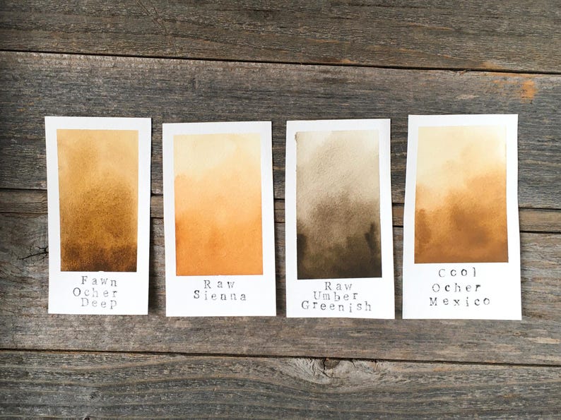 May include: Four rectangular paper swatches displaying watercolor gradients. The labels read Fawn Ocher Deep, Raw Sienna, Raw Umber Greenish, and Cool Ocher Mexico. Each swatch features a smooth transition from a light tone at the top to a darker, saturated hue at the bottom, showcasing distinct earth-toned pigments.