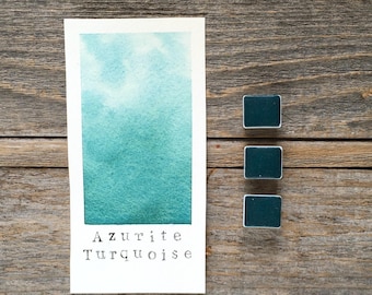 May include: A rectangular paper swatch featuring a watercolour gradient from light to dark teal, labelled Azurite Turquoise in black text. Beside the swatch are three small, square, dark teal watercolour pans arranged in a vertical column.