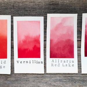 Op de afbeelding: Vier aquarelverfstaaltjes op een houten oppervlak. De staaltjes zijn gelabeld met "Marigold Orange", "Vermilion", "Alizarin Red Lake" en "Rose Madder".