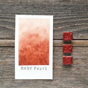 May include: A rectangular watercolor swatch card featuring a gradient of Ruby Pearl paint, transitioning from a light peach to a deep metallic red, with the text Ruby Pearl printed below. Three small square pans of metallic red watercolor paint are included.