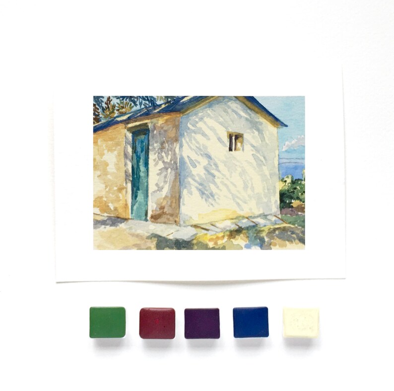 May include: A watercolor painting of a small building with a blue door and a white wall featuring tree shadow patterns. Below the painting are five square watercolor paint pans in green, maroon, purple, blue, and off-white colors.
