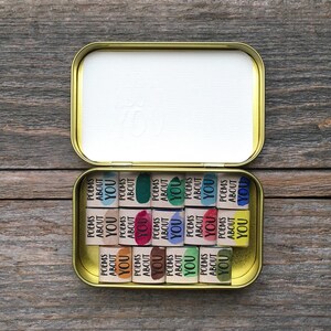May include: A gold-colored metal tin containing fifteen small rectangular blocks. Each block features the printed text "POEMS ABOUT YOU" alongside a unique colored paint swatch, including shades of blue, green, pink, yellow, and brown.