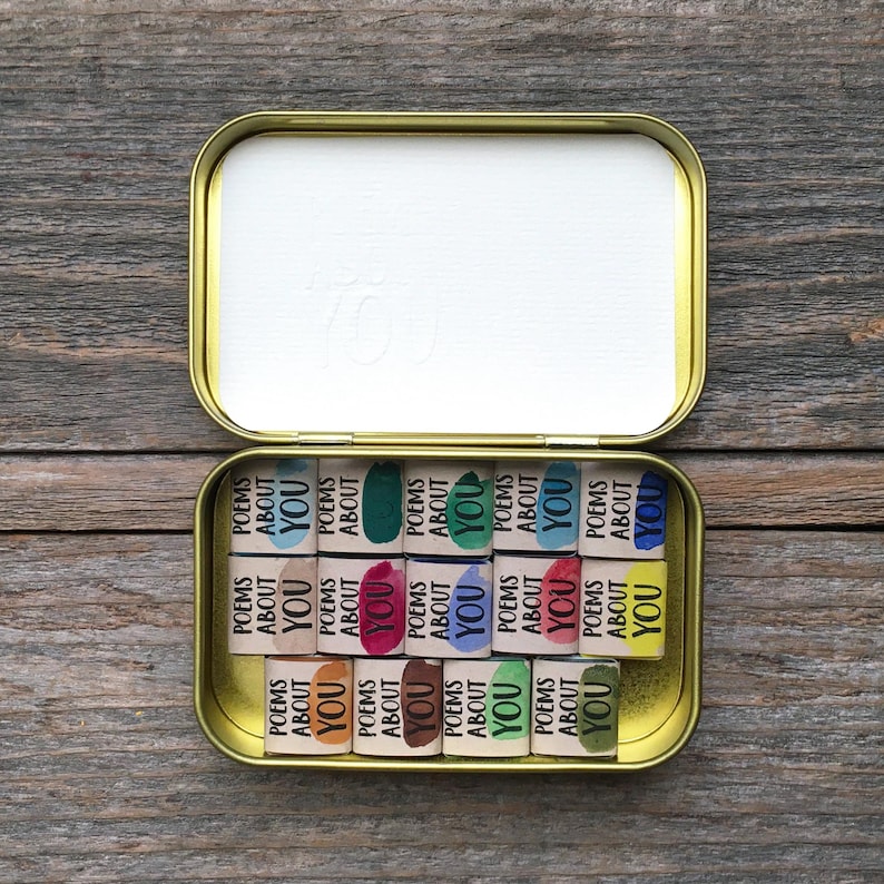 May include: A gold-colored metal tin containing fifteen small rectangular blocks. Each block features the printed text "POEMS ABOUT YOU" alongside a unique colored paint swatch, including shades of blue, green, pink, yellow, and brown.