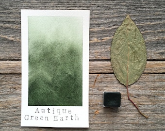 May include: A watercolor paint swatch in a green shade labeled "Antique Green Earth". A dried leaf and a small black paint pan are also shown on a wooden surface.