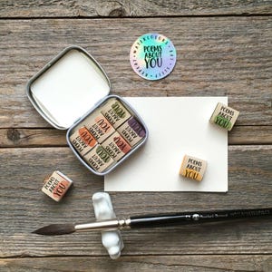May include: A set of small square watercolor pans in a metal tin, each labeled POEMS ABOUT YOU. Includes a round holographic sticker with the text WATERCOLORS EAT SLEEP REPEAT POEMS ABOUT YOU, a blank white card, and a black paintbrush.