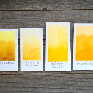 May include: Four rectangular paper swatches displaying watercolor gradients in shades of yellow. Each swatch is labeled with black text: Gamboge, Indian Yellow, Dalamar Yellow, and Diarylide. The colors transition from light to deep yellow tones.
