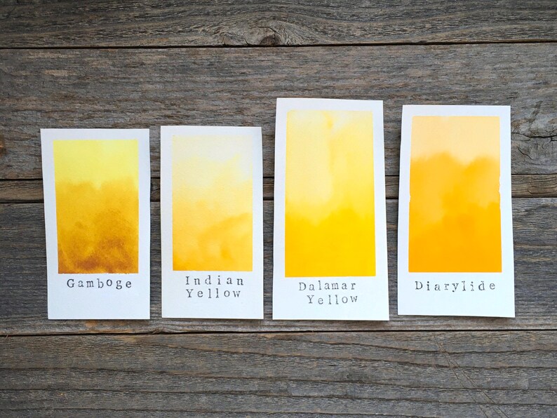 May include: Four rectangular paper swatches displaying watercolor gradients in shades of yellow. Each swatch is labeled with black text: Gamboge, Indian Yellow, Dalamar Yellow, and Diarylide. The colors transition from light to deep yellow tones.