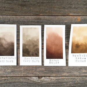 May include: Four rectangular paper swatches displaying watercolor paint gradients in shades of brown. The labels read Burnt Umber Very Dark, Raw Umber Very Dark, Burnt Umber, and Buckskin Brown Ocher, showing color transitions from light to dark.