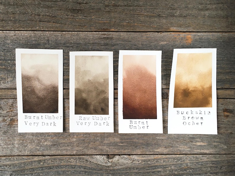 May include: Four rectangular paper swatches displaying watercolor paint gradients in shades of brown. The labels read Burnt Umber Very Dark, Raw Umber Very Dark, Burnt Umber, and Buckskin Brown Ocher, showing color transitions from light to dark.