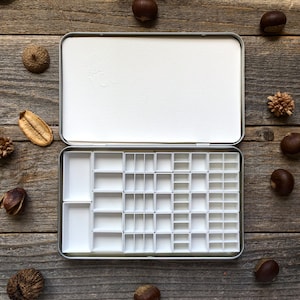 Może przedstawiać: Metalowa paleta do farb akwarelowych w kolorze srebrnym z 36 pustymi otworami. Paleta jest otwarta i leży na drewnianej powierzchni.