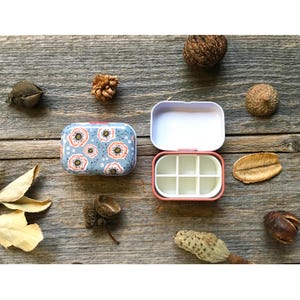 May include: Two rectangular, floral-patterned pill boxes, one open and one closed, sit on a rustic wooden surface. The closed box features a blue background with white and orange flower designs. The open box reveals a pink interior with six compartments.