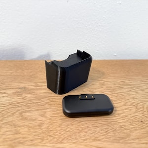May include: A black charging dock and a small, rectangular black device with gold-coloured contact points. The charging dock has a curved design with a slot for the device. The items are on a wooden surface.