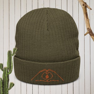 May include: A green knitted beanie with a fold-over cuff. The beanie has an orange embroidered design of two hands forming a heart shape with an Om symbol inside.