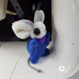 Könnte beinhalten: Ein handgefertigtes Plüschmaus-Spielzeug mit blauem Körper, weißen und grauen Ohren und großen schwarzen Augen. Die Maus hat einen lila Schal und einen grauen Schnur-Schwanz. Das Spielzeug liegt auf einer weißen Oberfläche.
