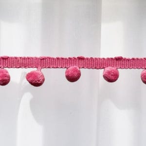 May include: A strip of dark pink pom-pom trim featuring a woven fabric header and a series of small, round, fuzzy spheres attached along the bottom edge. The trim is presented in a horizontal orientation, showcasing the consistent spacing of the pom-poms.