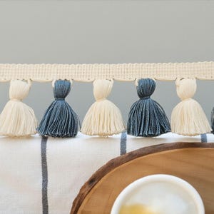 May include: Cream-coloured trim with alternating navy blue and cream tassels. The trim is above a white and blue striped fabric, with a wooden tray and a white cup in the foreground. Decorative home decor.