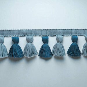 Puede incluir: Un ribete decorativo con una fila de borlas azules y grises alternas. Las borlas están suspendidas de una banda tejida azul claro. Adecuado para proyectos de artesanía y diseño.